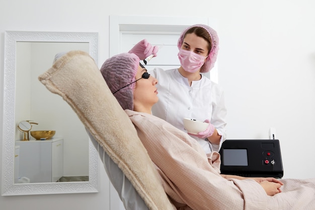 Carbon peeling in beauty salon cosmetologist applying black mask on the face of a beautiful woman