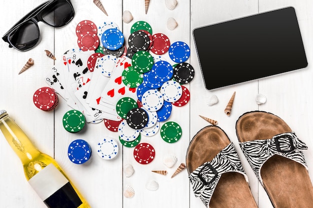 Card deck surrounded by poker chips and scattered seashells, glasses, a tablet and bottle of beer, on white wooden background with copy space