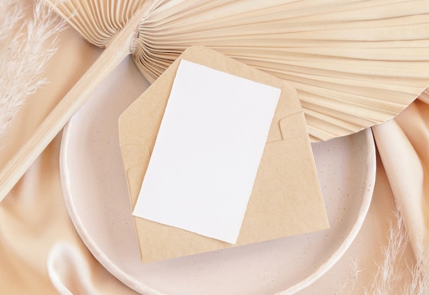 Card and envelope on plate on dried palm leaf and beige silky fabric close up greeting or wedding mockup