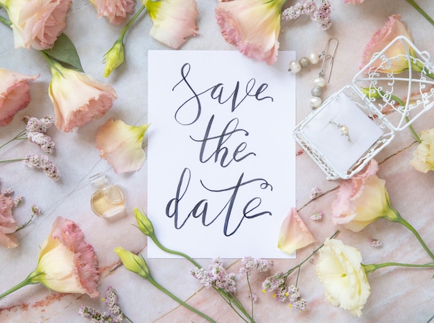 Card with the handwritten text SAVE THE DATE surrounded by pink flowers, earrings, petals and parfume flacon top view on a marble table. Romantic concept