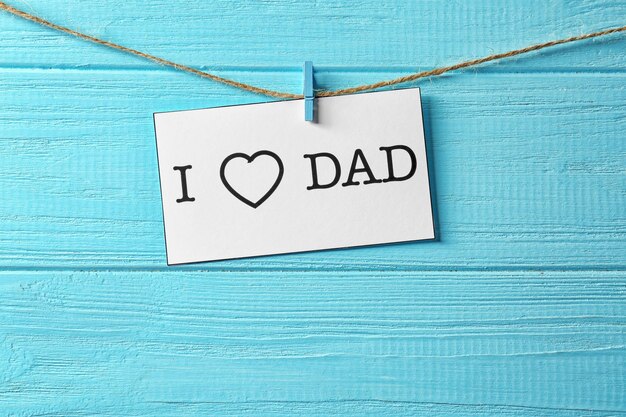 Card with text I LOVE DAD hanging on rope against wooden background