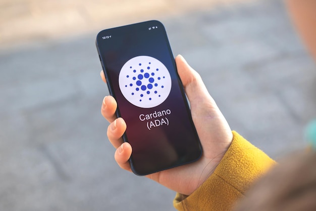 Cardano ADA cryptocurrency-symbool, logo. Zakelijk en financieel concept. Hand met smartphone, scherm met crypto icoon close-up