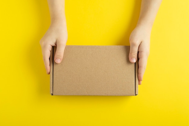 Cardboard box in child hands on yellow background, top view.