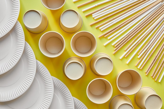 Cardboard straws in the upper right corner a row of cardboard cups in the center and some white cardboard plates in the lower left corner of the image on a yellow background