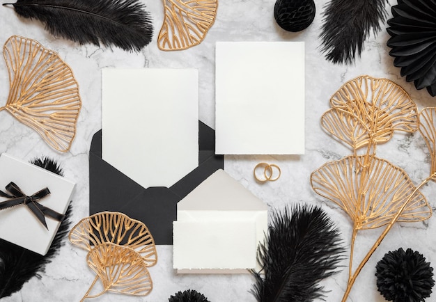 Cards and envelope on marble table near black feathers and golden leaves Wedding mockup