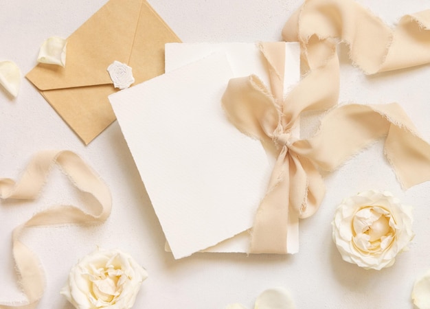 Фото Карты, привязанные бежевой шелковой лентой рядом с кремовыми розами и конвертом на белом макете стола
