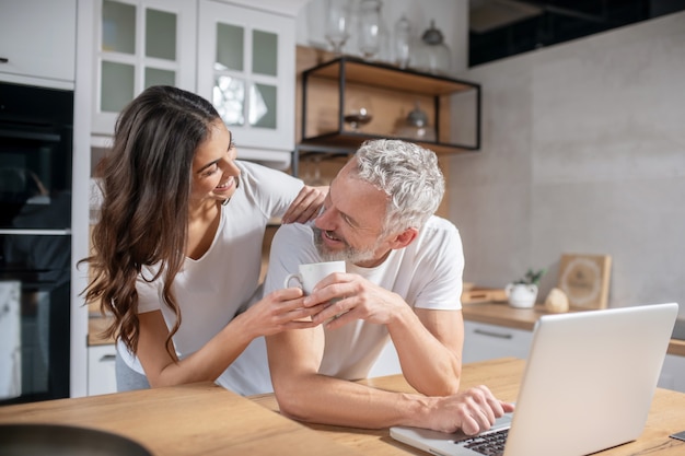 Care. Caring joyful long haired wife giving coffee to smiling gray haired husband working at desk with laptop