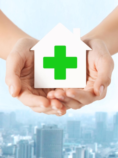 care, help, charity and people concept - close up of hands holding white paper house with green cross sign over city background