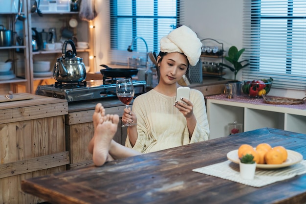 Carefree asian female with towel on head and legs on dining\
table is reading social media feeds on the smartphone and enjoying\
red wine after bath at home.