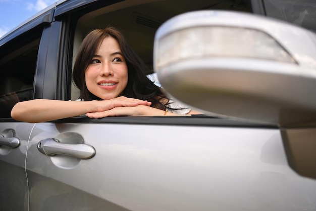 Carefree asian woman leaning out of a car window and enjoying nature during summer road trip