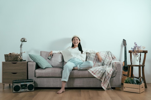 беззаботная азиатская женщина смотрит телевизор дома, сидя на диване в современной зимней квартире. Молодая смеющаяся случайная девушка наслаждается телешоу, держа пульт дистанционного управления. Комфортный образ жизни copyspace