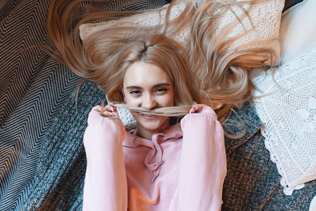 Carefree beautiful girl playing with her long blond hair while lying on the bed at home.