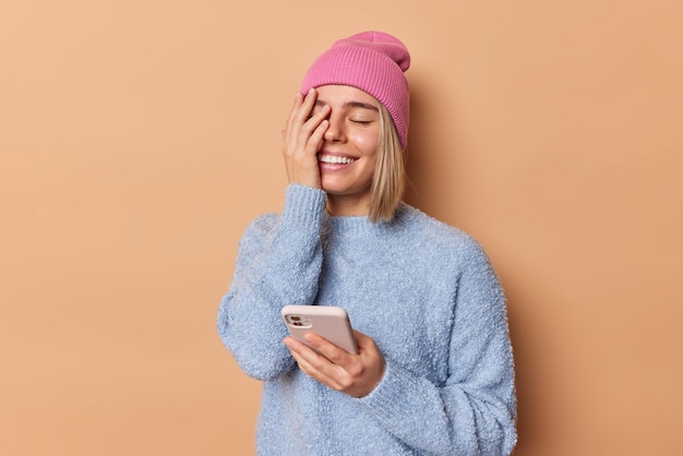 평온하고 쾌활한 귀여운 여성은 얼굴을 손바닥으로 웃게 만들고 온라인 채팅을 위해 휴대폰을 기쁘게 사용하며 분홍색 모자를 쓰고 베이지색 배경 위에 격리된 점퍼는 남자 친구로부터 즐거운 메시지를 받습니다.