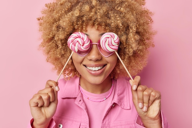 Фото Беззаботная жизнерадостная женщина закрывает глаза карамельными спиральными конфетами, позитивно хихикает, одетая в куртку, изолированную на розовом фоне, без ума от сладостей люди и концепция сахарной зависимости