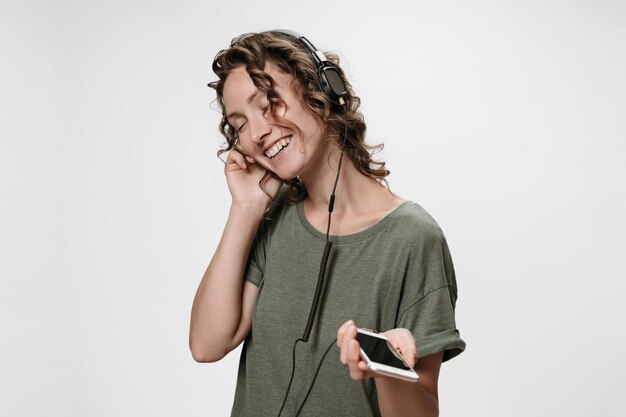 Carefree cheerful young curly woman listen favourite music with\
her stereo headphones, satisfied with quality of sound, enjoy\
favourite playlist, smiles, being in high spirit, isolated on\
white.