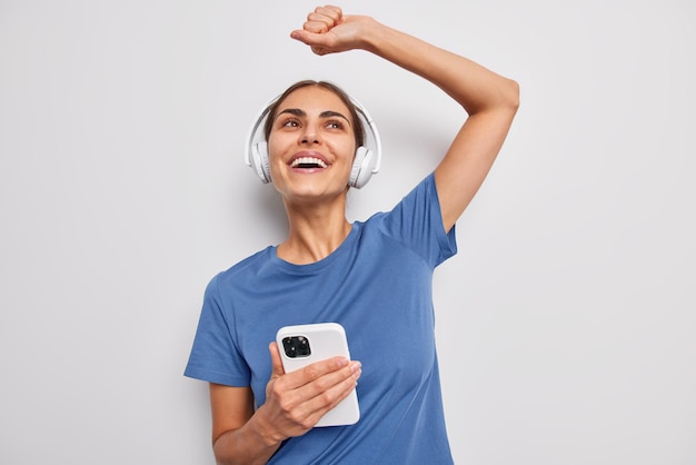 Carefree cheerful young woman dances with arm raised listens
music vis wireless headphones in app wears casual blue t shirt has
upbeat mood isolated over white background enjoys favorite
songs