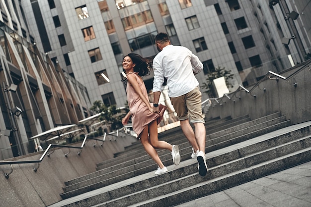 Carefree day together. Full length rear view of playful young couple holding hands and smiling while running up the stairs outdoors