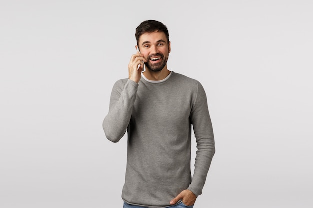 Carefree enthusiastic, bearded male in grey sweater talking on phone with pleased, happy smile, hold hand in pockets, use smartphone calling via internet messanger