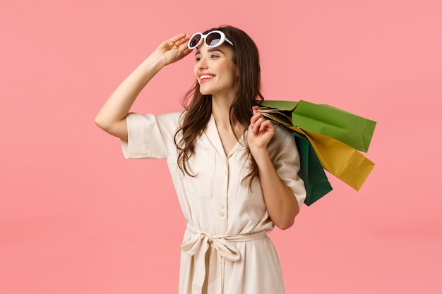 Carefree enthusiastic woman in dress, taking off glasses and looking happily sideways as enjoying beautiful day, fantastic shopping with discounts, holding shop bags behind back, pink wall