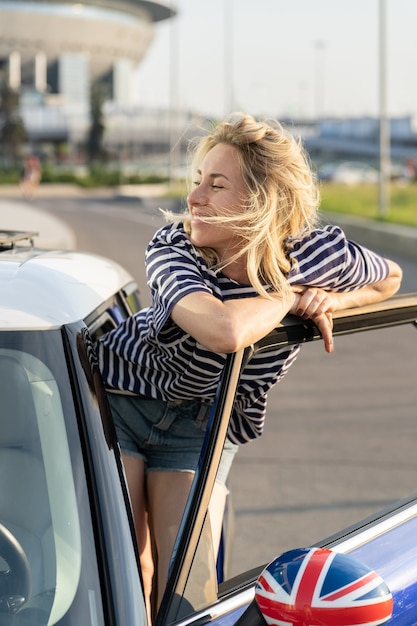 열린 자동차 문에서 자신감 있는 운전사를 둔 평온한 여성은 휴가를 위해 여행하기 전에 신선한 공기를 즐깁니다