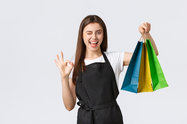 Carefree friendly-looking female cashier in black apron guarantee quality or purchased items, wink and show okay sign while handing over paper bags with customer order, white background