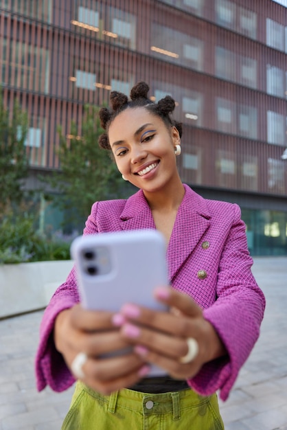 Carefree glad millennial girl with trendy hairstyle makes selfie photo on smartphone smiles positively wears fashionable outfit poses against urban background during daytime strolls in city.