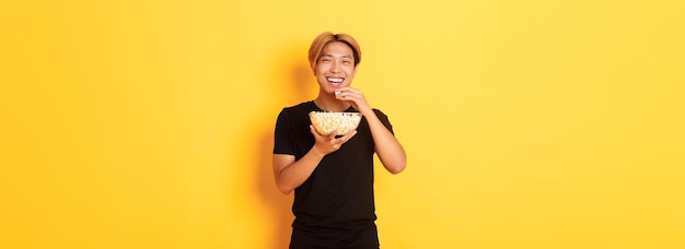 금발 머리를 가진 평온한 행복한 아시아 남자는 코미디가 웃고 노란색으로 서서 팝콘을 먹는 것을 보고 있습니다.