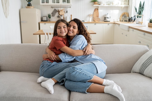 Carefree happy family of millennial woman and teenage girl hugging sits on big sofa in living room