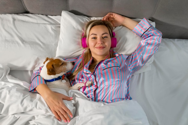 Беззаботная дама в пижаме слушает музыку в наушниках, сидя в постели со своим любимым питомцем