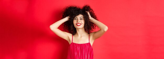 Carefree lady in red dress touching her curly hair and smiling happy standing on studio background