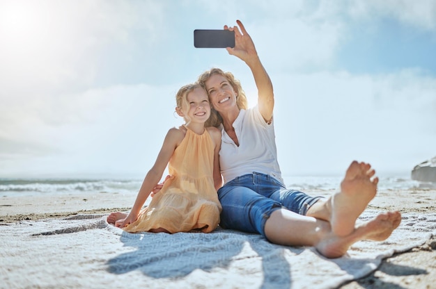 ビーチに座って自分撮りをしているのんきな母と娘休日に携帯電話で写真を撮っている間笑顔の幸せな少女と祖母ママと娘の絆