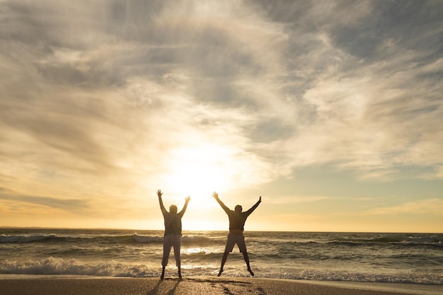 Carefree multiracial senior couple jumping with arms raised at beach against sky during sunset