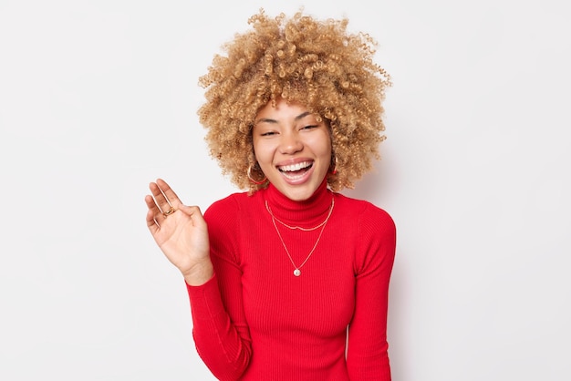 写真 巻き毛の笑顔でのんきな楽観的な女性は、白い背景の上に分離された赤いタートルネックに身を包んだ面白いジョークで、手のひらを上げて喜んで笑い続けます。人と感情の概念
