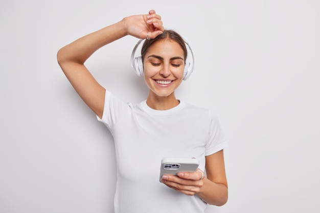 Photo carefree positive teenage girl moves with rhythm of music holds mobile phone wears wireless headphones on ears dressed casually isolated over white wall