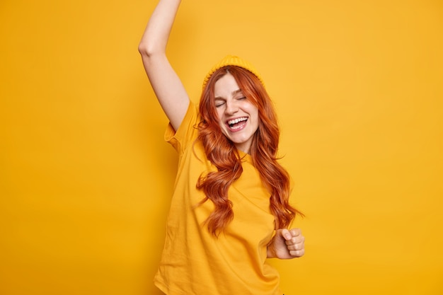 Carefree positive young ginger girl dances happily keeps arm raised closes eyes feels upbeat dressed in casual t shirt and hat poses against vivid yellow wall has fun enjoys successful day