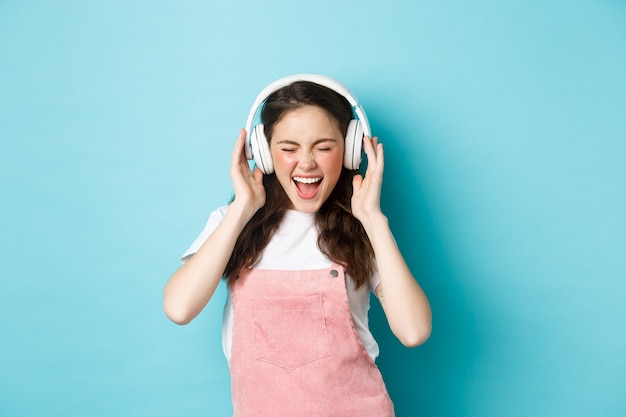 Carefree pretty girl scream from joy, listening music in headphones with happy face, lip-sync lyrics, standing over blue background.