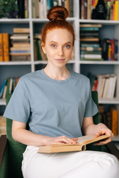 La studentessa spensierata della giovane donna rossa sta leggendo il libro gode del riposo nell'ufficio di casa che guarda l'obbiettivo signora carina che si gode i libri nella stanza della biblioteca sullo sfondo degli scaffali