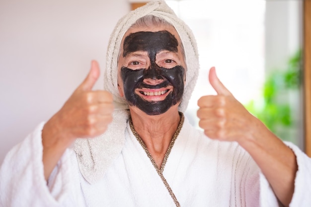 Carefree senior beautiful woman with a detox facial charcoal mask homemade smiling looking at camera with thumb up take care of the skin concept