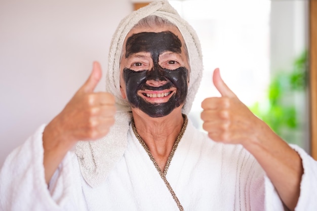 La bella donna anziana spensierata con una maschera di carbone facciale disintossicante sorridente fatta in casa guardando la fotocamera con il pollice in su si prende cura del concetto di pelle