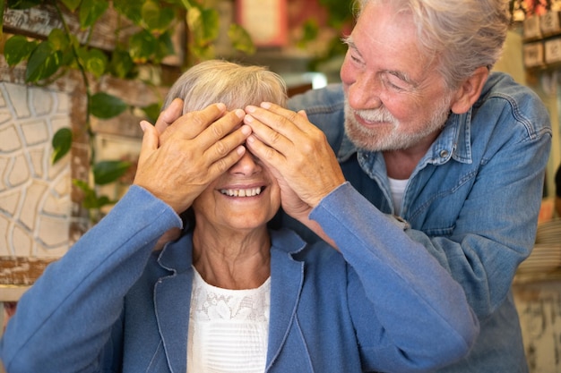 평온한 노인 부부는 커피숍에서 쾌활하게 웃고 있습니다. 손으로 여자의 눈을 덮고 있는 남자