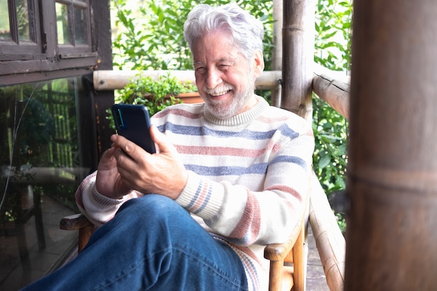 Carefree senior man sitting in wooden balcony of mountain chalet using phone. Bearded caucasian elderly grandfather relaxing in mountain and nature