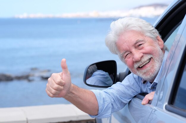 Carefree smiling adult whitehaired senior man sitting inside the car with hand out of the window and thumb up looking at camera Elderly grandparent with white beard enjoying travel Horizon over water