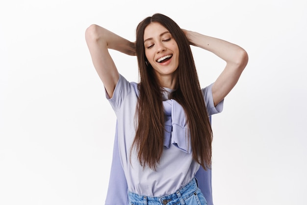 Carefree smiling brunette woman, resting, enjoying spring weekend, holding hands behind head and relaxing, looking aside, standing over white wall