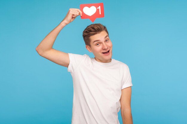 Carefree smiling man in white t-shirt holding social media button over his head, showing heart like icon, giving recommendation to support content, follow blog. indoor studio shot blue background