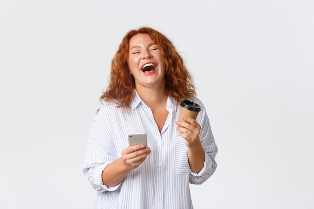 Беззаботная улыбающаяся женщина средних лет искренне смеется, держа чашку с кофе из кафе и мобильного телефона