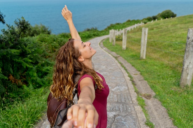 Carefree woman reaching hand to camera in green pathway. Horizontal view of backpacker on adventure trip. People and travel destination concept.