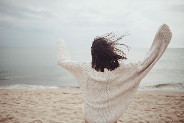 冷たい海の砂浜を走る風の強い髪のセーターを着た屈託のない女性の後ろ姿