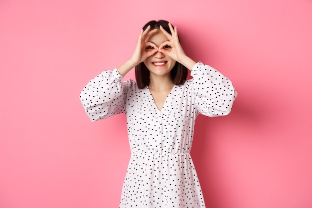 Carefree young asian woman looking through hand binoculars at camera, staring at discounts, standing over pink background