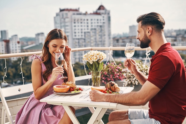 Carefree young couple in casual clothing enjoying romantic dinner and smiling while sitting on the rooftop patio outdoors