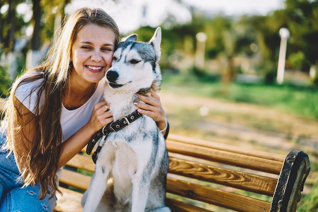 Carefree young female student stroking friendly grey husky and resting in summer park